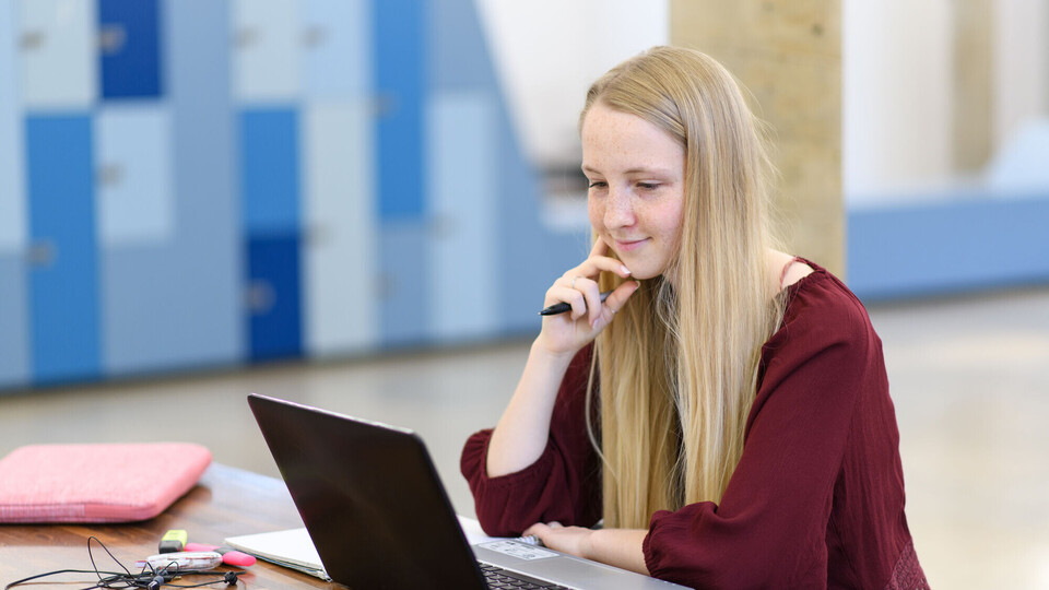 Eine Studentin sitzt mit ihrem Laptop an einem Tisch und betrachtet etwas auf dem Bilschirm.