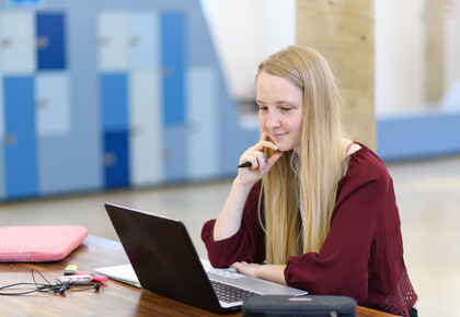 Eine Studentin sitzt mit ihrem Laptop an einem Tisch und betrachtet etwas auf dem Bilschirm.
