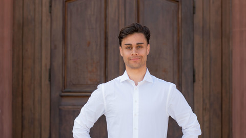 Lucas Currado. He is wearing a white shirt and is standing in front of a brown door.