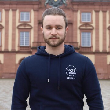 Magnus hat dunkelblonde Haare und Bart. Außerdem hat er einen dunkelblauen Kapuzenpullover an. Er schaut freundlich in die Kamera.