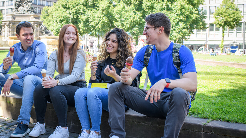Vier Studierende sitzen im Stadtzentrum auf einer Mauer und essen Eis. Sie schauen sich an und lachen dabei. Im Hintergrund sind eine Wiese, Bäume und Gebäude zu sehen.