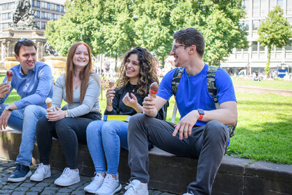 Vier Studierende sitzen im Stadtzentrum auf einer Mauer und essen Eis. Sie schauen sich an und lachen dabei. Im Hintergrund sind eine Wiese, Bäume und Gebäude zu sehen.