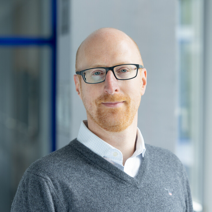 Florian Keusch hat eine Glatze und einen rothaarigen Bart. Er trägt eine schwarz umrandete Brille und einen grauen Wollpullover.