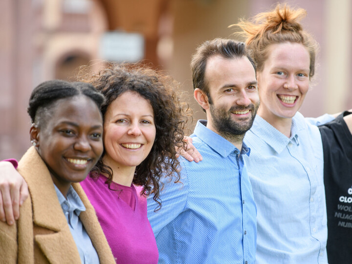 4 Personen haben die Arme jeweils auf die Schultern des*der Nachbarperson gelegt.