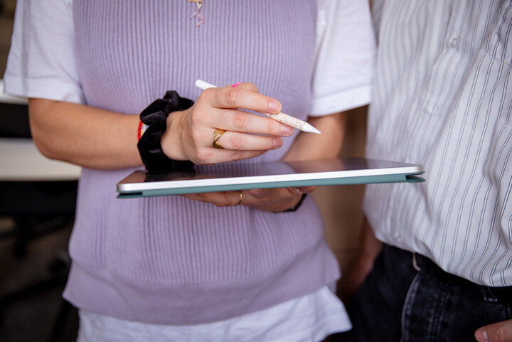 Bildausschnitt eines Oberkörpers einer Person mit fliederfarbenem Pullunder. In der einen Hand hält sie ein Tablet, in der anderen einen Stift.
