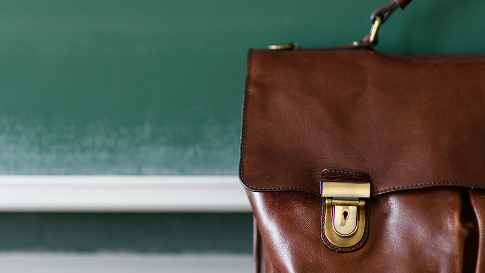 Eine braune Ledertasche steht im Vordergrund einer Kreidetafel