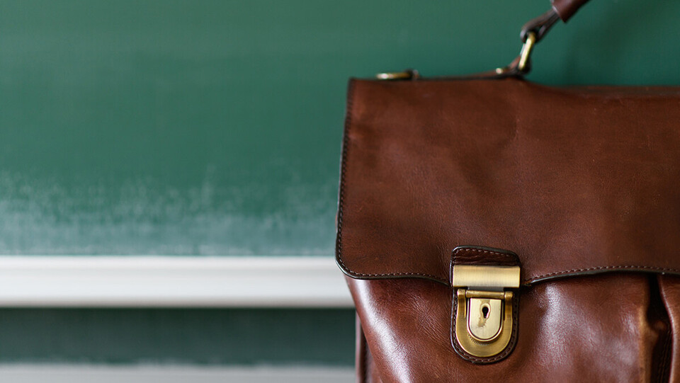 Eine braune Ledertasche steht im Vordergrund einer Kreidetafel