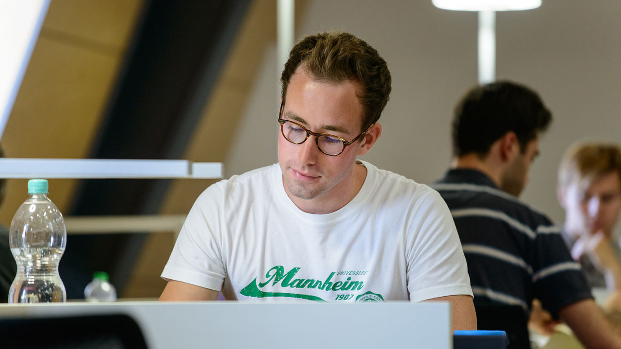 Ein Student lernt an einem Tisch. Er trägt kurzes braunes Haar, eine runde Brille und ein weißes T-Shirt mit der grünen Aufschrift "Mannheim".