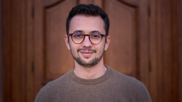 Alexander Gnaedinger. He is wearing a beige sweater and is standing in front of one the castle doors.
