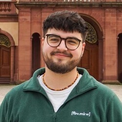 Daniel Wiggenhauser hat braune Haare und einen Vollbart. Er trägt einen grünen Pullover und  trägt eine Brille.