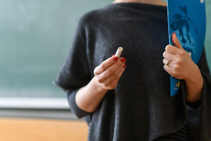 Eine Frau im schwarzen T-Shirt steht vor einer grünen Kreidetafel und hält ein stück Kreide in der manikürten Hand und ein Heft unterm Arm. Eine Lehrerin?