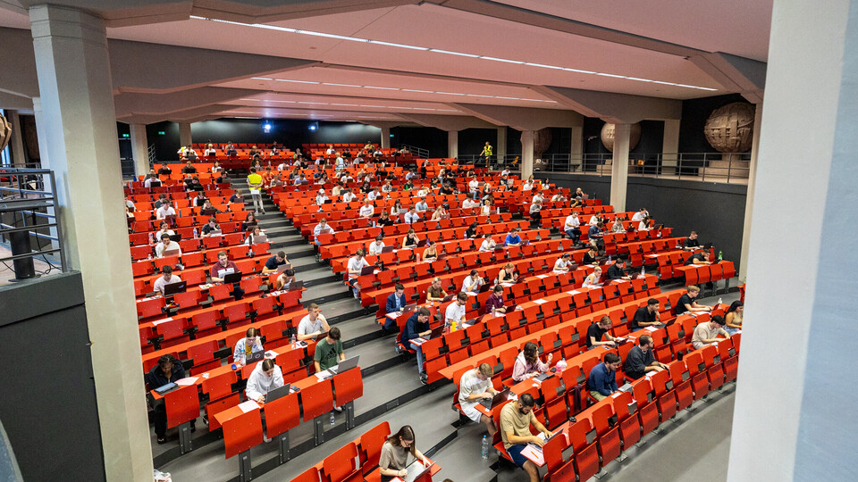 Studierende schreiben eine digitale Prüfung mit Laptops in einem großen Hörsaal mit roten Sitzreihen unter Aufsicht.