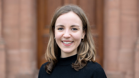 Lena Rudat. She is wearing a black sweater and is standing in front of a brown door and wall.