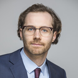 Paul Beck hat braune Haare und einen Dreitagebart. Er trägt eine Brille und einen blauen Blazer mit Krawatte.