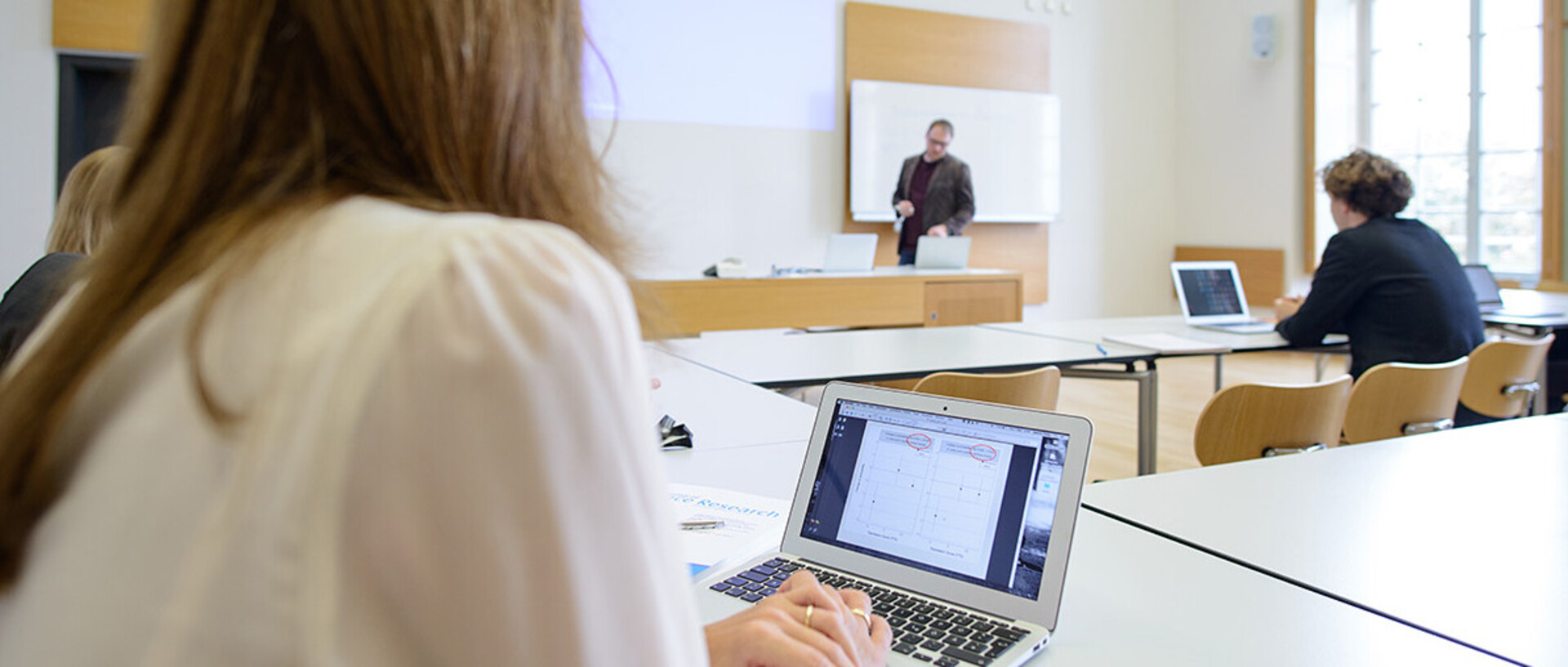 Studierende in einem Seminarraum, am Laptop, eine Lehrperson steht an der Tafel und erklärt