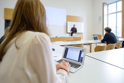 Studierende in einem Seminarraum, am Laptop, eine Lehrperson steht an der Tafel und erklärt