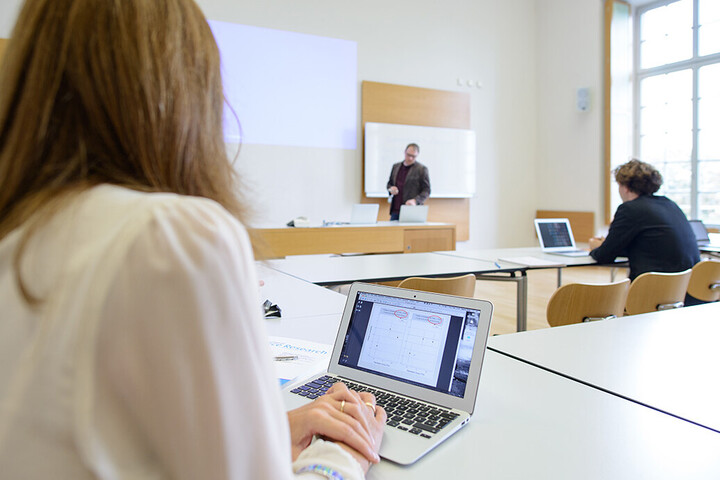 Studierende in einem Seminarraum, am Laptop, eine Lehrperson steht an der Tafel und erklärt