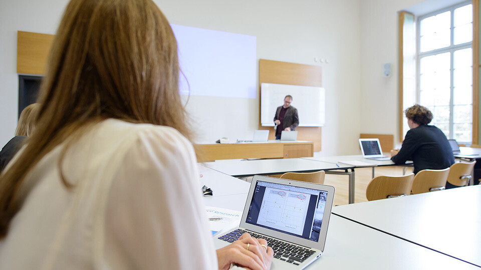 Studierende in einem Seminarraum, am Laptop, eine Lehrperson steht an der Tafel und erklärt