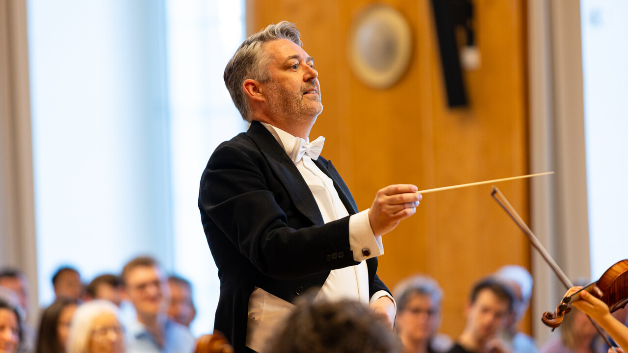 Auftritt des Uniorchesters. Dirgent trägt einen schwarzen Anzug und steht vor dem Publikum.