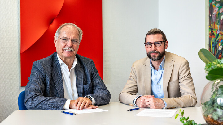 Dr. Peter Merten und Christian Haas sitzen an einem Tisch. Sie schauen in die Kamera. Vor ihnen liegen Zettel.