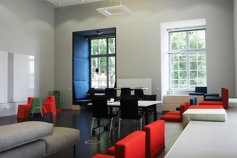 In einem hellen Raum der Bibliothek im Schneckenhof liegen bunte Sitzkissen in rot und blau verteilt. An den großen Fenstern sind gemütliche Ecken.