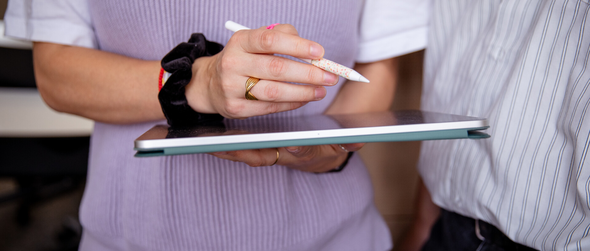 Eine weiblich gelesenen Personen hält ein Tablet in den Händen und arbeiten mit einem Digital-Pen daran. Eine zweite Person schaun ihr dabei zu. Die Personen sind nicht vollständig zu erkennen.
