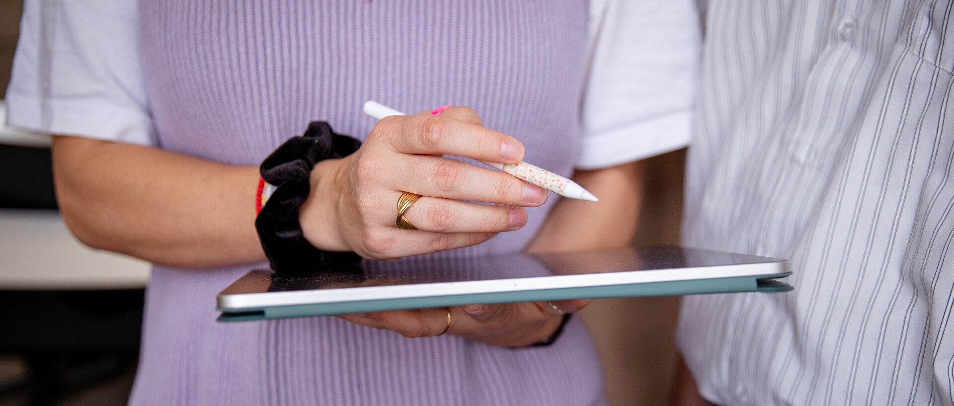 Eine weiblich gelesenen Personen hält ein Tablet in den Händen und arbeiten mit einem Digital-Pen daran. Eine zweite Person schaun ihr dabei zu. Die Personen sind nicht vollständig zu erkennen.