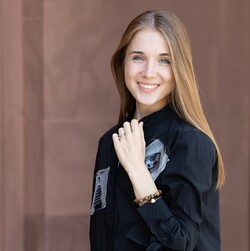 Eine lächelnde Person trägt ein schwarzes Kleid und steht vor einer braunen Steinwand. Die Person heißt Alena Shmalko.