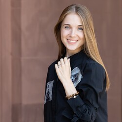 Eine lächelnde Person trägt ein schwarzes Kleid und steht vor einer braunen Steinwand. Die Person heißt Alena Shmalko.