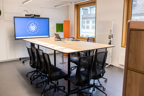Das Bild zeigt einen Konferenztisch für zwölf Personen. Im Hintergrund ist ein großes Display mit dem Logo der Universität Mannheim zu sehen.
