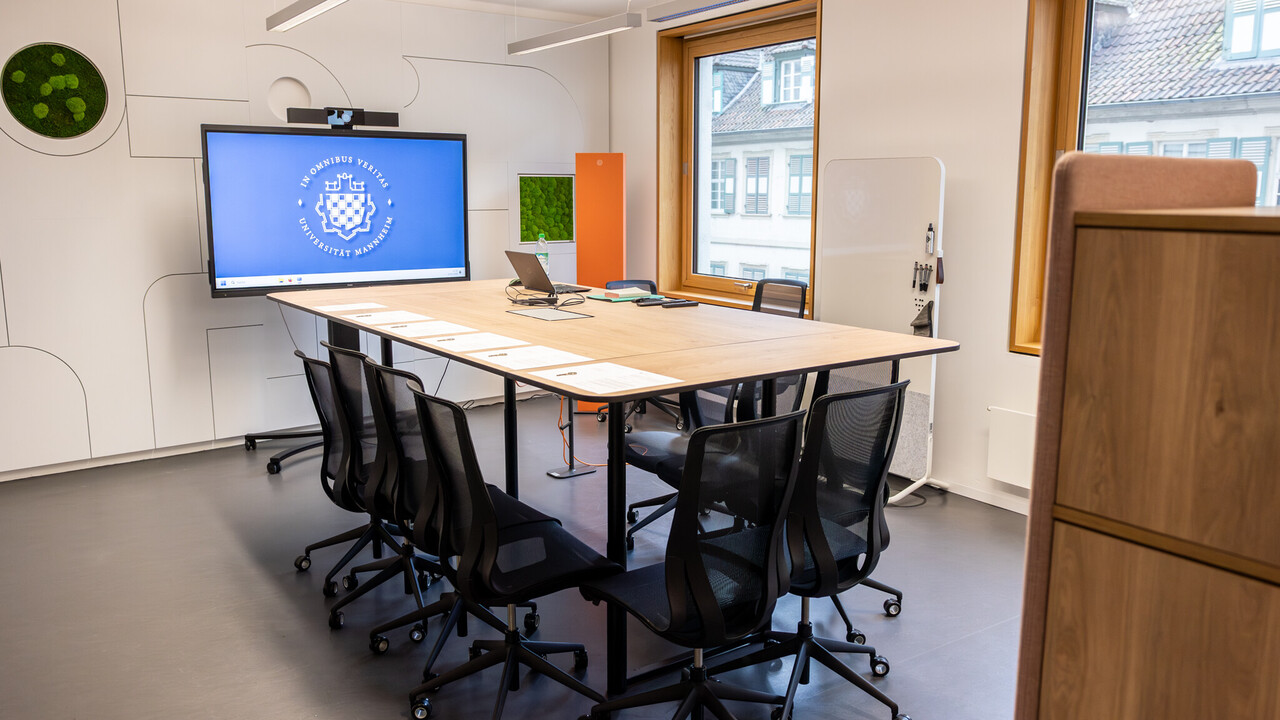Das Bild zeigt einen Konferenztisch für zwölf Personen. Im Hintergrund ist ein großes Display mit dem Logo der Universität Mannheim zu sehen.
