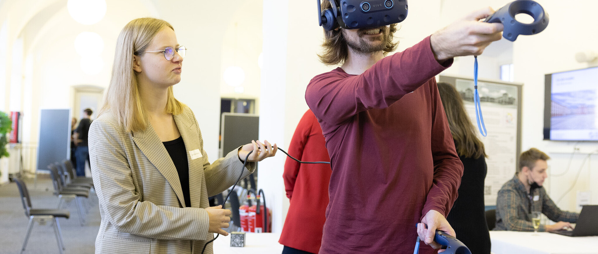 Ein Mann mit VR-Brille bewegt sich im Raum. Neben ihm eine Frau, die das Kabel der Brille hält. 