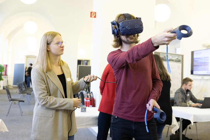 Ein Mann mit VR-Brille bewegt sich im Raum. Neben ihm eine Frau, die das Kabel der Brille hält. 