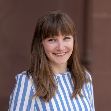 Caterina Katzer hat braune lange Haare und ein Pony. Sie trägt ein blau-weiß-gestreiftes Oberteil.