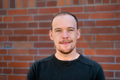 Jan Menzner. He is wearing a black shirt and is standing in front of a brickwall.