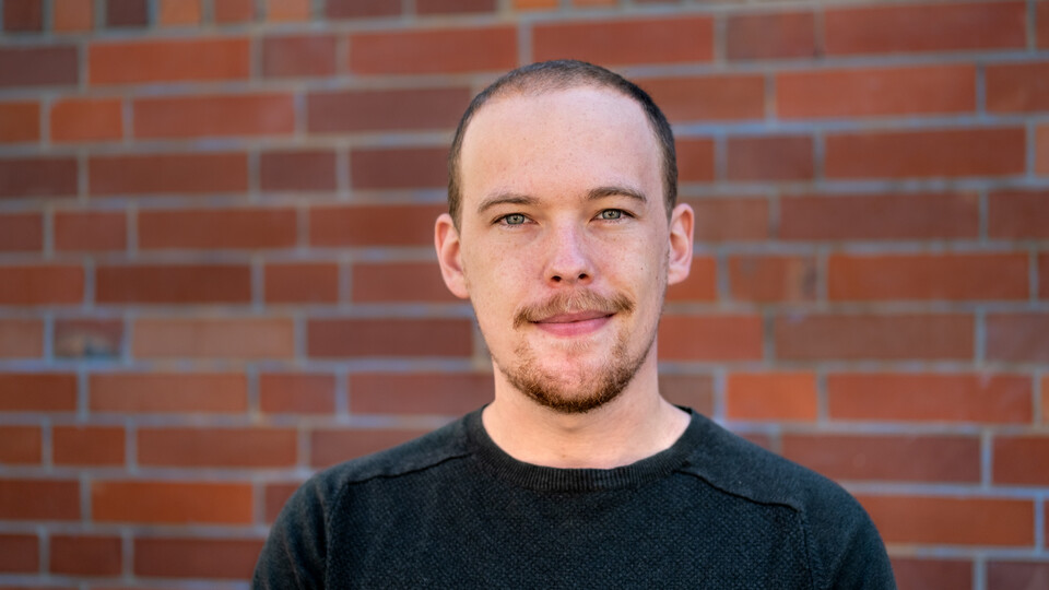 Jan Menzner. He is wearing a black shirt and is standing in front of a brickwall.