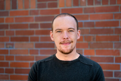 Portrait of Jan Menzner. He is wearing a black sweater and standing in front of a wall.
