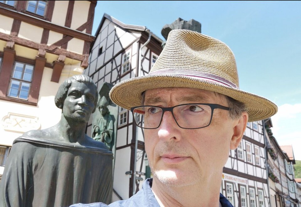Mann mit Hut und Brille vor einer Statue und Fachwerkhäusern