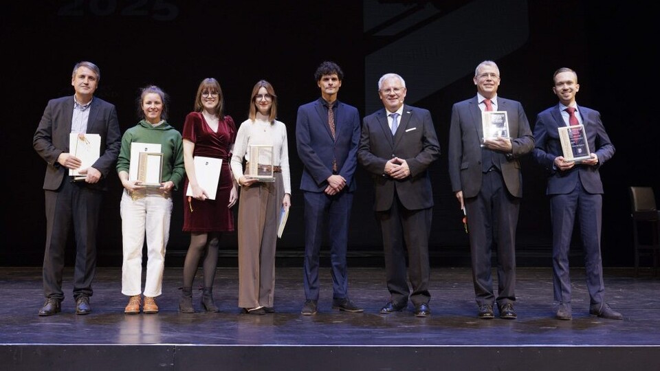 Auf einer Bühne vor schwarzem Hintergrund stehen nebeneinander die Preisträger*innen (drei Frauen und fünf Männer) in schicker Kleidung. 