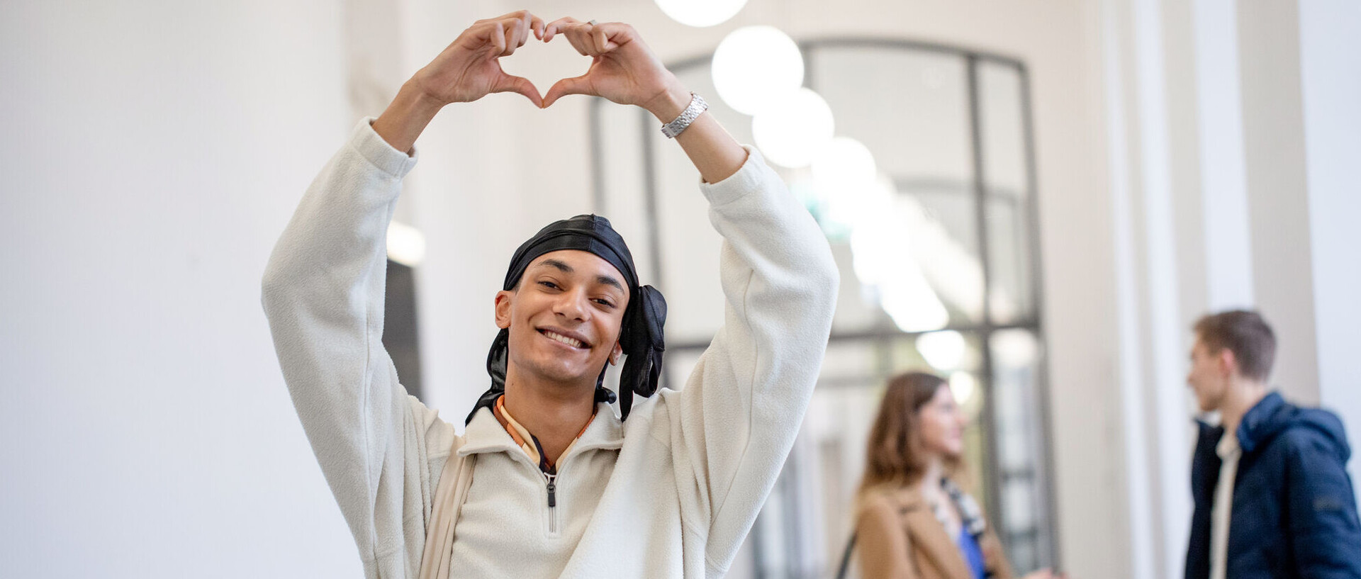StudyCheck Award 2026 Ein Student steht im Flur des Schlosses und lächelt in die Kamera. Er hält seine Arme nach oben und formt mit den Händen ein Herz. Im Hintergrund unterhalten sich eine Studentin und ein Student.