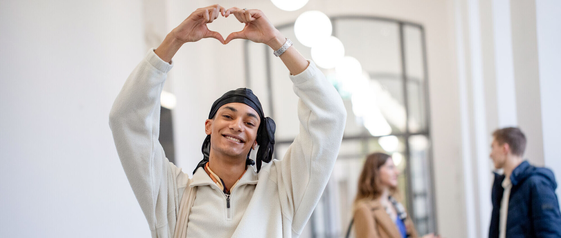 Ein Student steht im Flur des Schlosses und lächelt in die Kamera. Er hält seine Arme nach oben und formt mit den Händen ein Herz. Im Hintergrund unterhalten sich eine Studentin und ein Student.