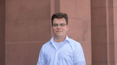 David Müller. He is wearing a white shirt and is standing in front of a brown wall.