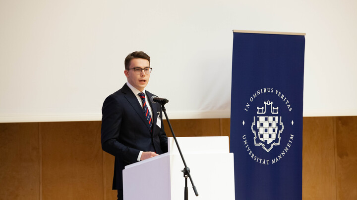 Eine Person trägt einen dunkelblauen Anzug und steht hinter einem Rednerpult. Neben dem Pult steht ein dunkelblaues Roll-Up der Uni Mannheim mit dem Logo der Uni Mannheim.