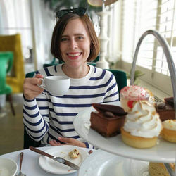 Eva Streng trägt einen braunen Bob, einen blau-weiß gestreiften Pullover und hält in ihrer Hand eine Tasse. Im Vordergrund steht eine Etagere mit Kleingebäck.