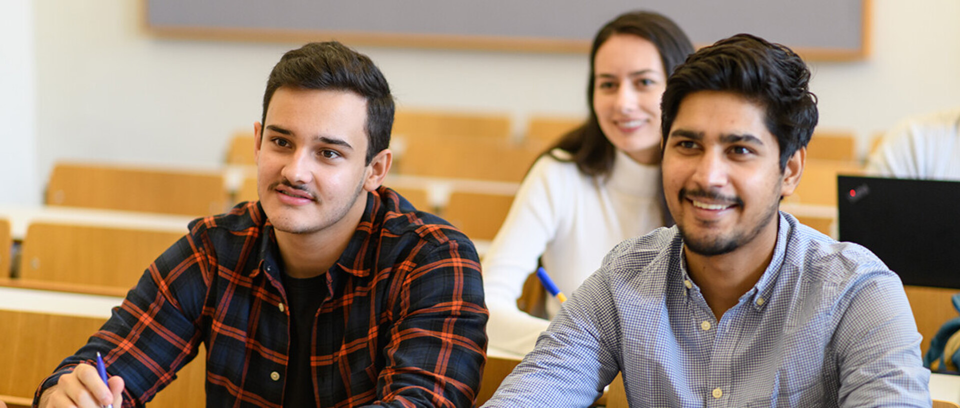 Zwei Studenten sitzen an einem Tisch in einem Seminarraum. Vor ihnen liegen Papier und Stifte.