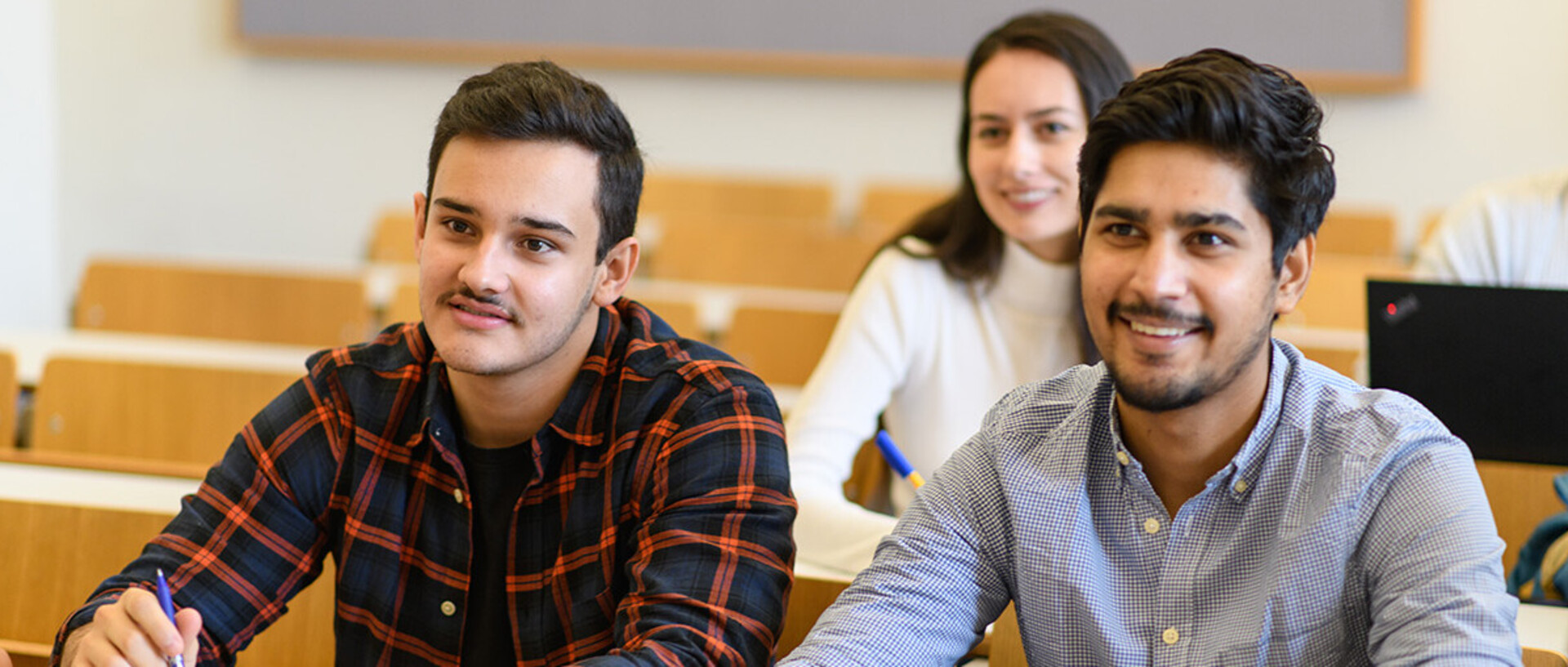 Zwei Studenten sitzen an einem Tisch in einem Seminarraum. Vor ihnen liegen Papier und Stifte.