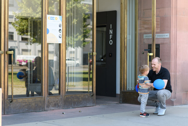 Ein Mitarbeitender der Universität Mannheim und sein junger Sohn lachen gemeinsam vor dem Haupteingang der Universität.