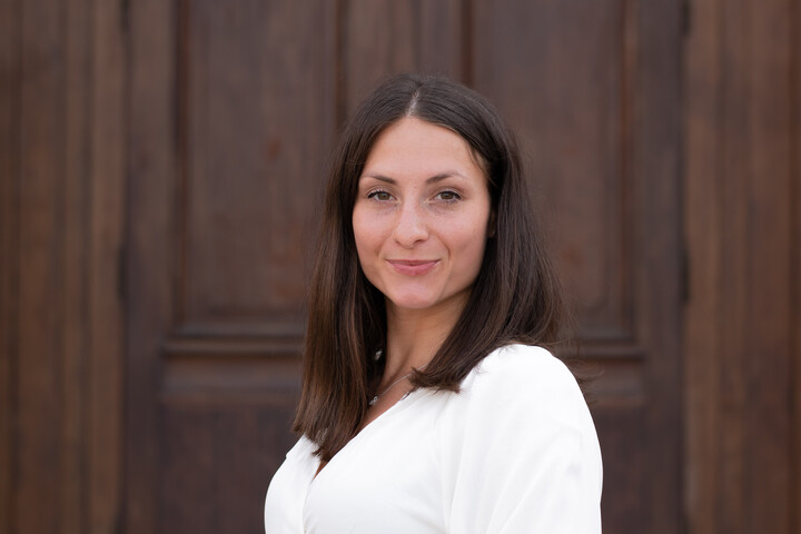 Giuliana Manganaro. Sie trägt eine weiße Bluse und steht vor einer Holztür.