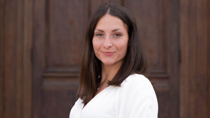 Giuliana Manganaro. She is wearing a white shirt and is standing in front of one of the castle doors.