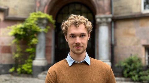 Max Otto. He is wearing a brown sweater over a white shirt and is standing in front a tall doorway arch.